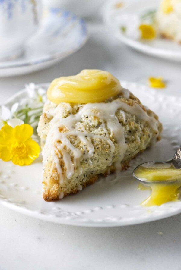 A poppyseed lemon scone on a plate with a spoonful of lemon curd and a tea cup in the background.