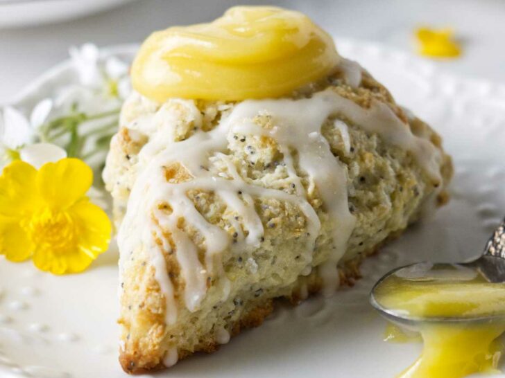 A poppyseed lemon scone on a plate with a spoonful of lemon curd and a tea cup in the background.
