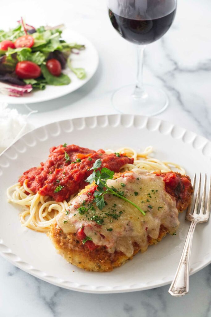 A serving of Olive Garden chicken parmesan with spaghetti and marinara sauce. Side salad and glass of wine in the background.