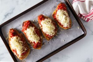 Four chicken breasts on a baking sheet topped with marinara sauce and cheese.