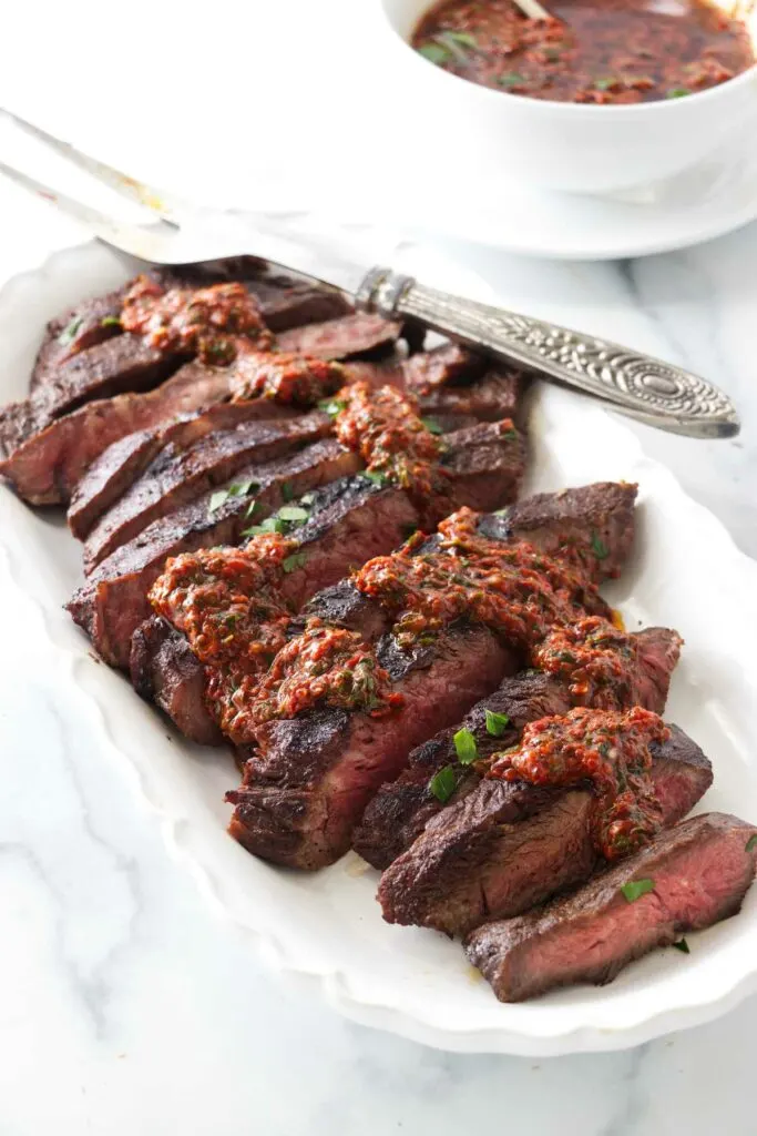 A platter of sliced sous vide flat iron steak and drizzled with red chimichurri sauce. A dish of red chimichurri sauce in the background.