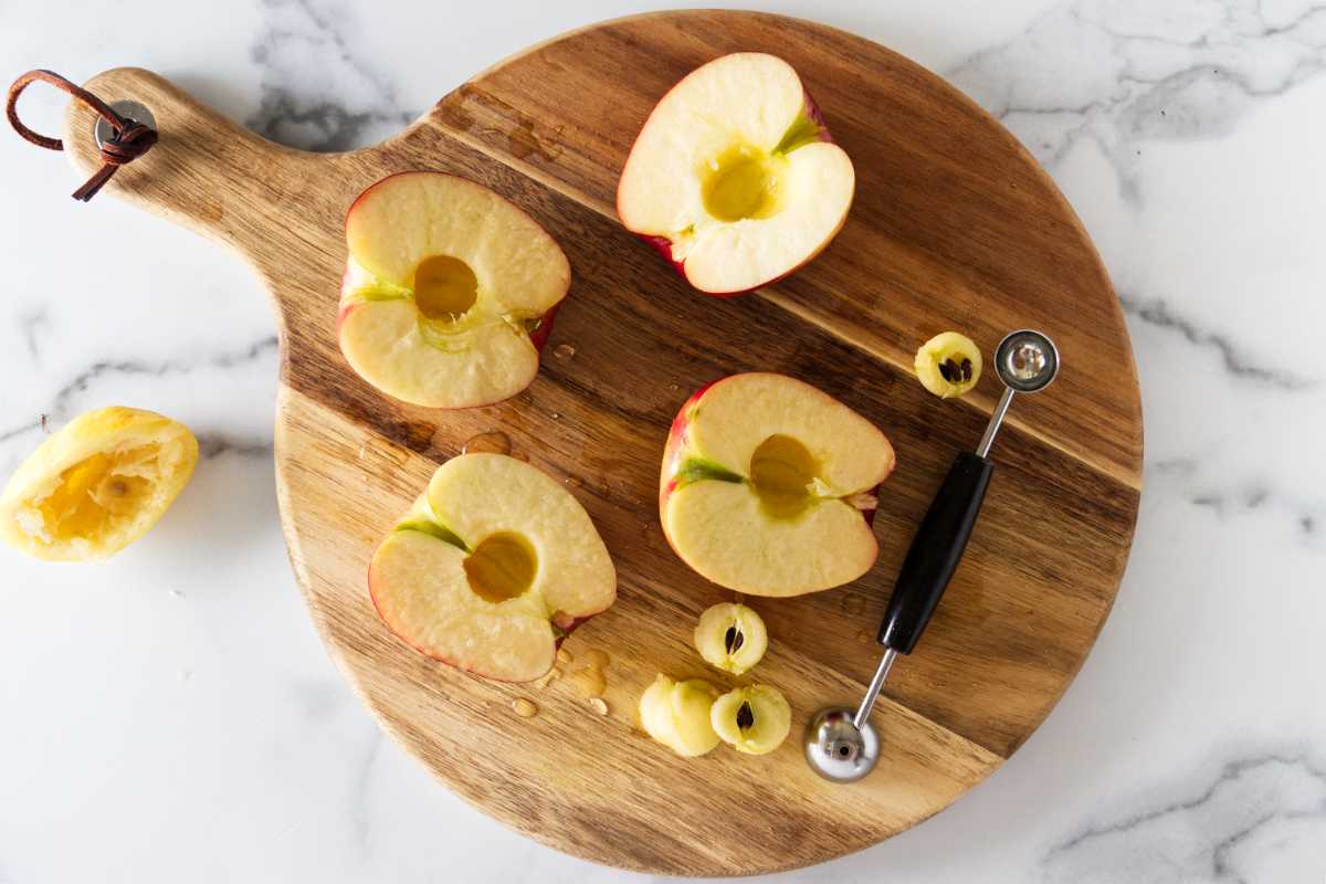 Large cutting board with halved and cored apples. Melon ball tool along side of the apples with rounds of core from the apples. A lemon rind to the left of the cutting board.