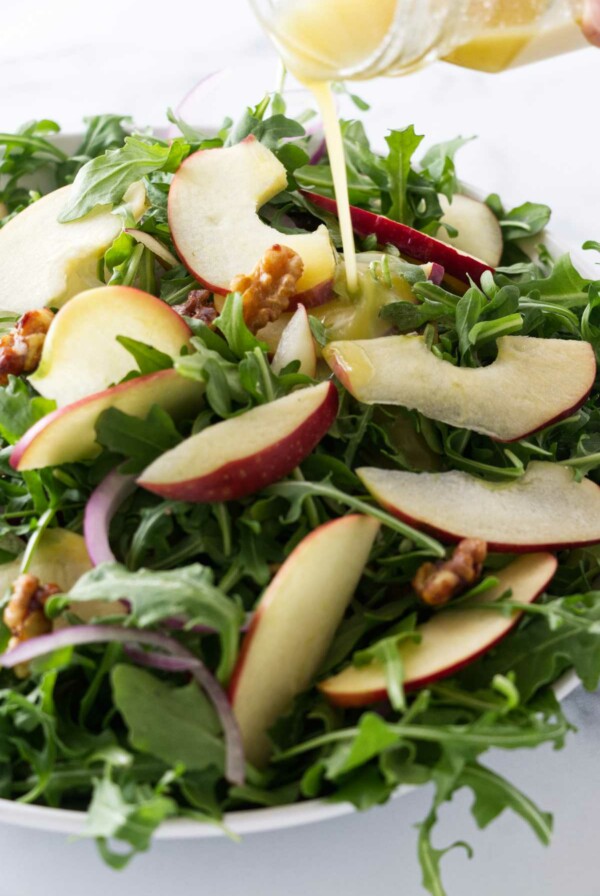 Salad bowl with arugula apple salad and a vinaigrette dressing being poured on.