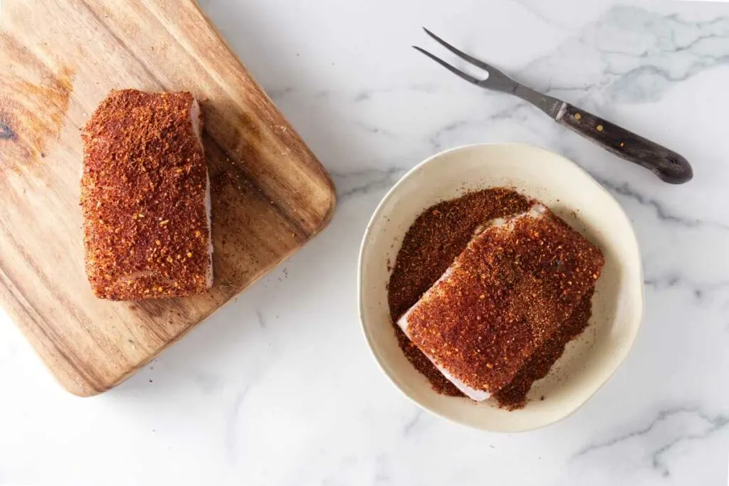 Halibut portions coated in blackening seasoning, on portion on a cutting board, one portion in a dish with the seasoning, fork alongside the dish.