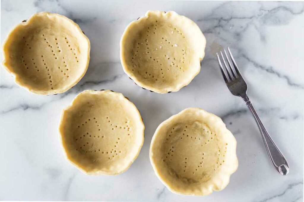 four docked pastry crusts in mini tart pans. A fork along side.