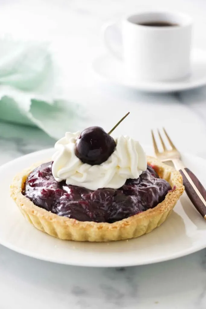 Mini cherry tart on a plate with a fork. Cup of coffee and napkin in the background.