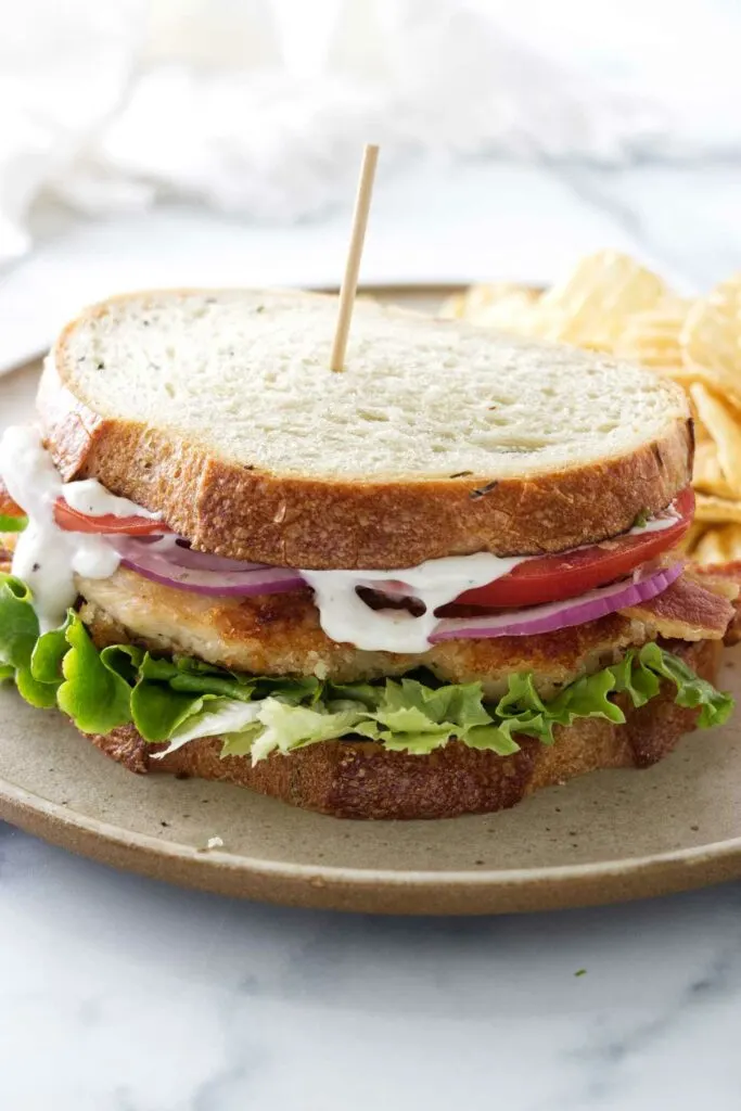 A crispy chicken bacon ranch sandwich on a plate with potato chips.