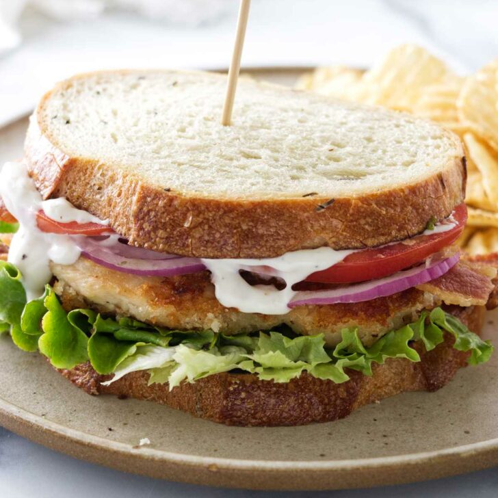 A crispy chicken bacon ranch sandwich on a plate with potato chips.