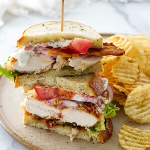 A plate with a chicken bacon ranch sandwich stacked and a wood food pick. Potato chips alongside the sandwich.