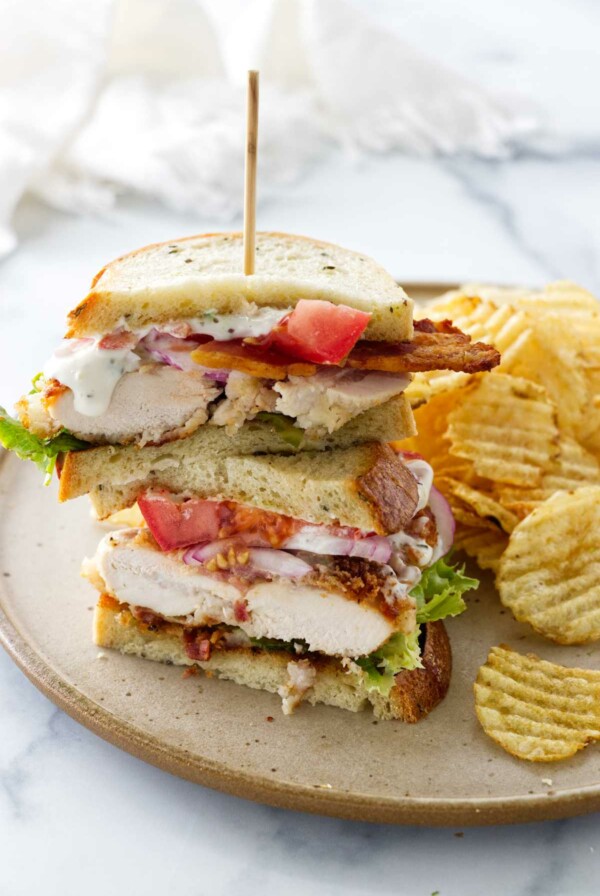 A plate with a chicken bacon ranch sandwich stacked and a wood food pick. Potato chips alongside the sandwich.