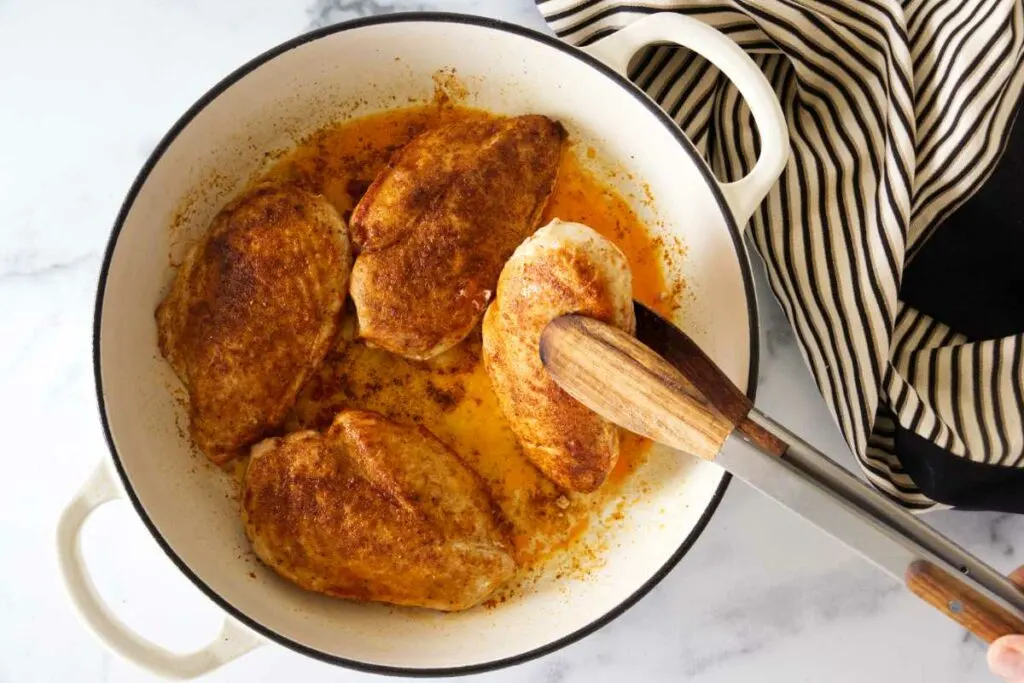 A pan with 4 seared paprika-seared chicken breasts.Tongs removing one chicken breast.