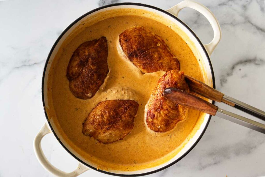 Tongs holding a serving of chicken breast and dding the seared chicken breasts back into the hot simmering paprika cream sauce.