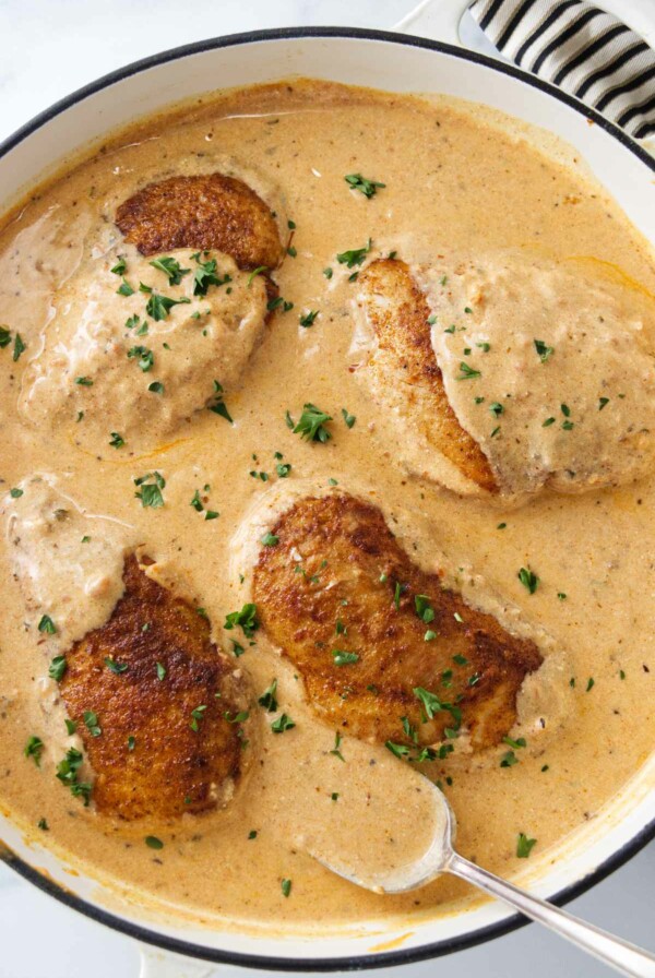 A pan of creamy paprika sauce and four chicken breasts. A spoon with sauce.