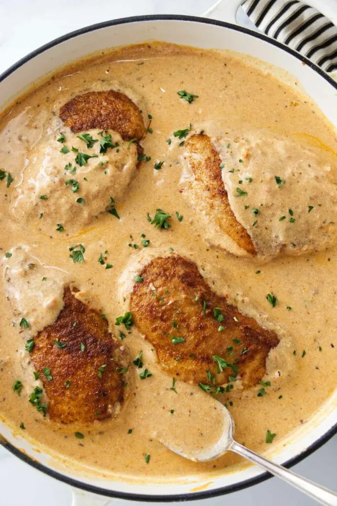 A pan of creamy paprika sauce and four chicken breasts. A spoon with sauce.