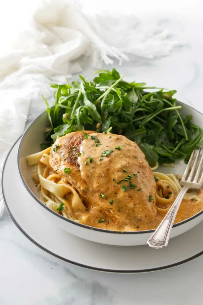 A serving of creamy paprika chicken on a bed of pasta with a salad and a fork. Napkin in the background.