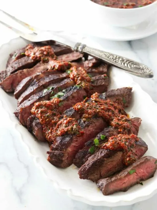 A platter of sliced sous vide flat iron steak and drizzled with red chimichurri sauce. A dish of red chimichurri sauce in the background.