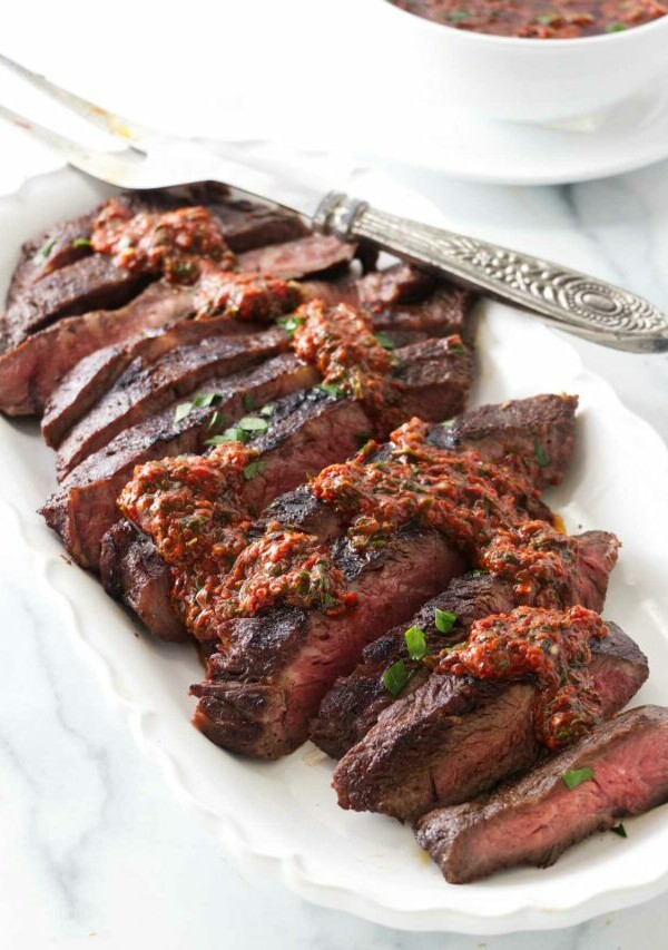 A platter of sliced sous vide flat iron steak and drizzled with red chimichurri sauce. A dish of red chimichurri sauce in the background.