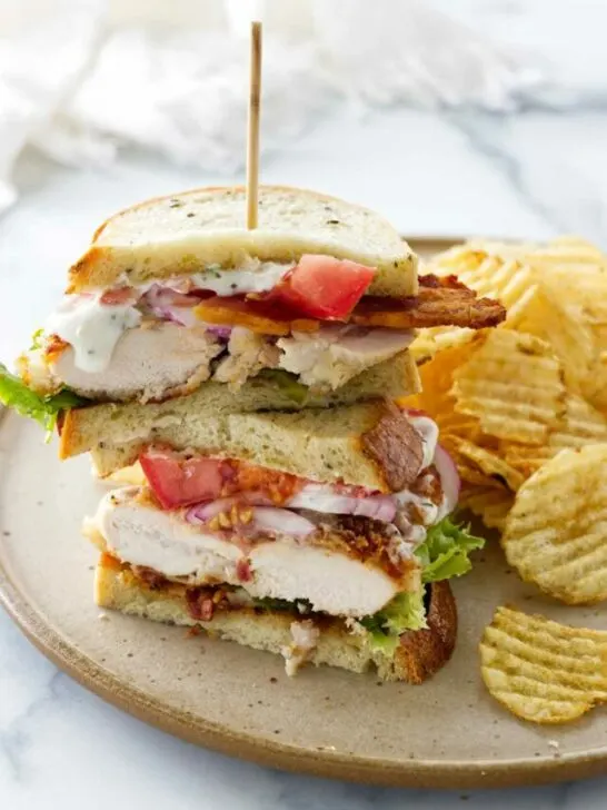 A plate with a chicken bacon ranch sandwich stacked and a wood food pick. Potato chips alongside the sandwich.