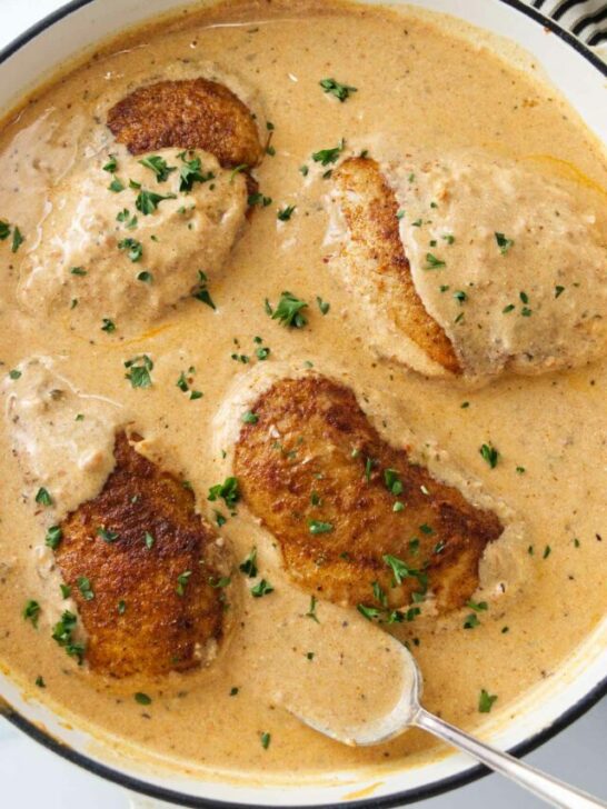 A pan of creamy paprika sauce and four chicken breasts. A spoon with sauce.