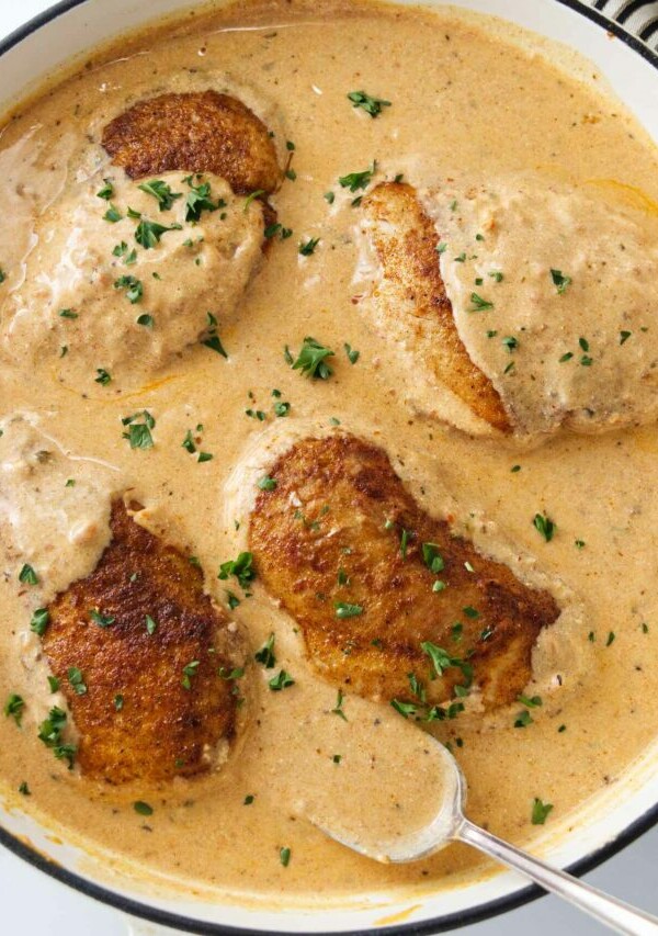 A pan of creamy paprika sauce and four chicken breasts. A spoon with sauce.