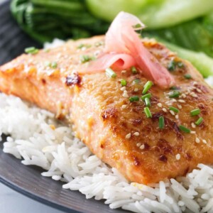 A plated serving of miso butter salmon on a mound of jasmine rice and garnished with sesame seeds, snipped fresh chives and pickled ginger. Bok choy in the background.