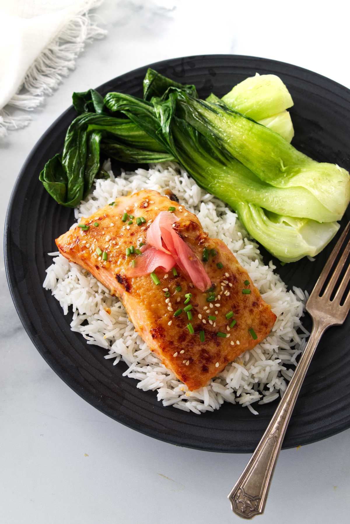 Overhead view of a plated serving of miso butter salmon on a mound of rice and steamed bok choy. A dinner fork on the plate and a napkin to the side.