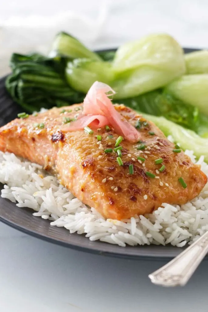 A plated serving of miso butter salmon on a mound of jasmine rice and garnished with sesame seeds, snipped fresh chives and pickled ginger. Bok choy in the background.