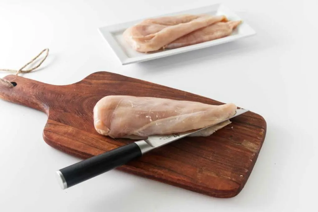 Chicken breast on a wood cutting board with a knife slicing it in half lengthwise. A small plate in the background with two chicken breasts.