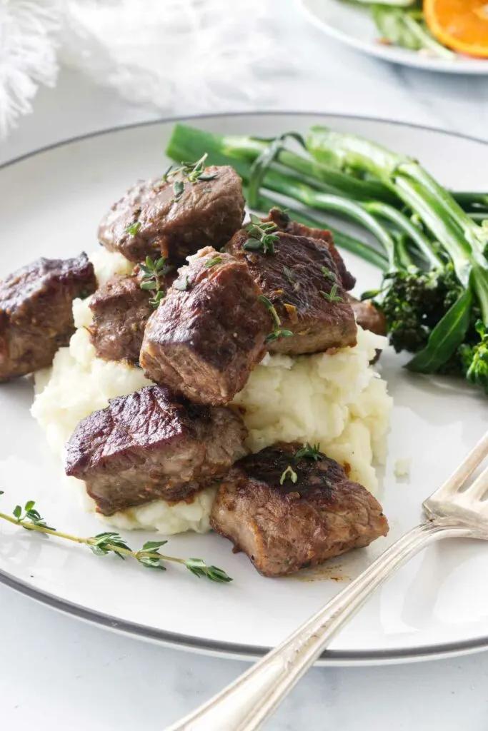 A serving of steak bites on a mound of mashed potatoes with broccolini and a dinner fork. A side salad in the background.