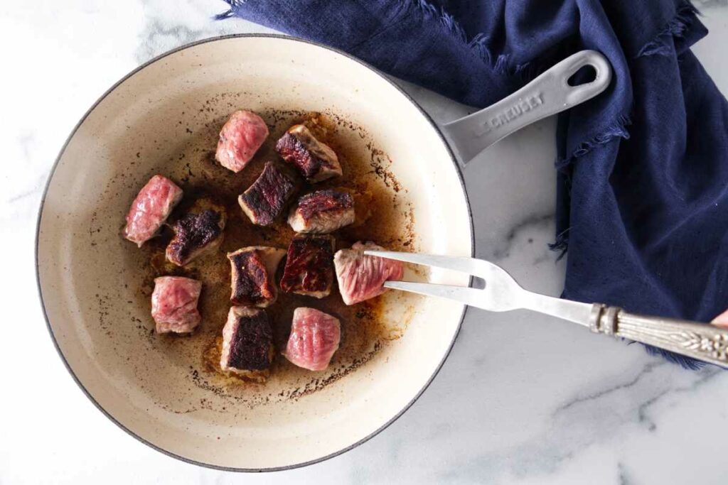 A skillet with a portion of steak bites being seared. A fork lifting one bite.