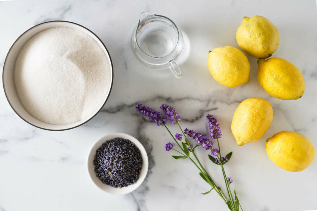 A bowl of sugar, pitcher of water, lemons, dried culinary lavender and fresh lavender blooms.