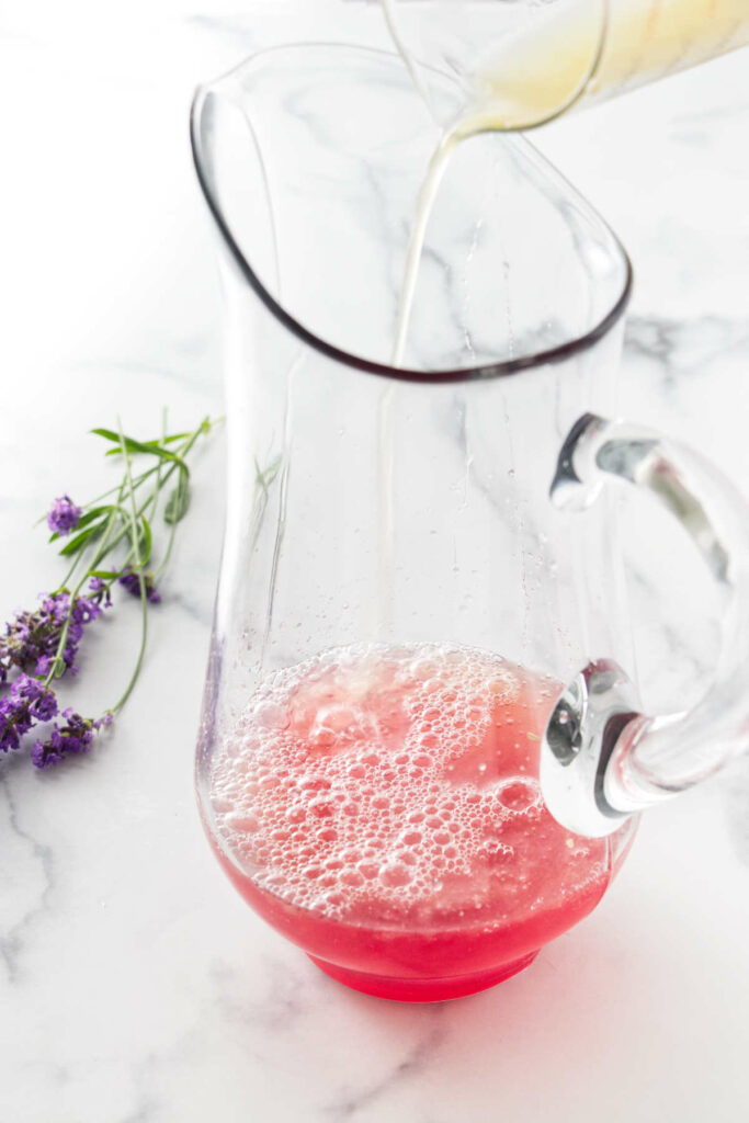 A clear glass pitcher with a pink juice and lemon juice being pored in it on a white marble background.