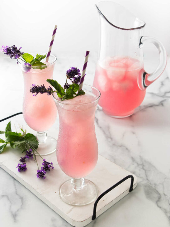 A clear glass pitcher and two tall glasses filled with a pink beverage sitting on a white tray with lavender blossoms.