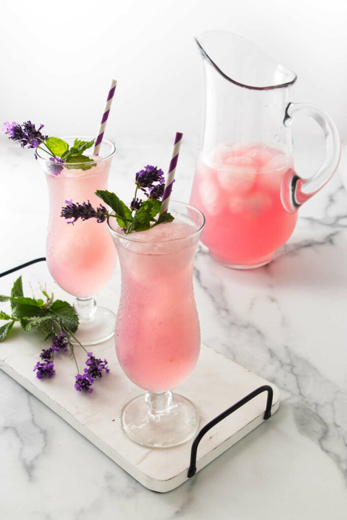 two glasses of lavender lemonade on a tray with a pitcher of lemonade in the background.