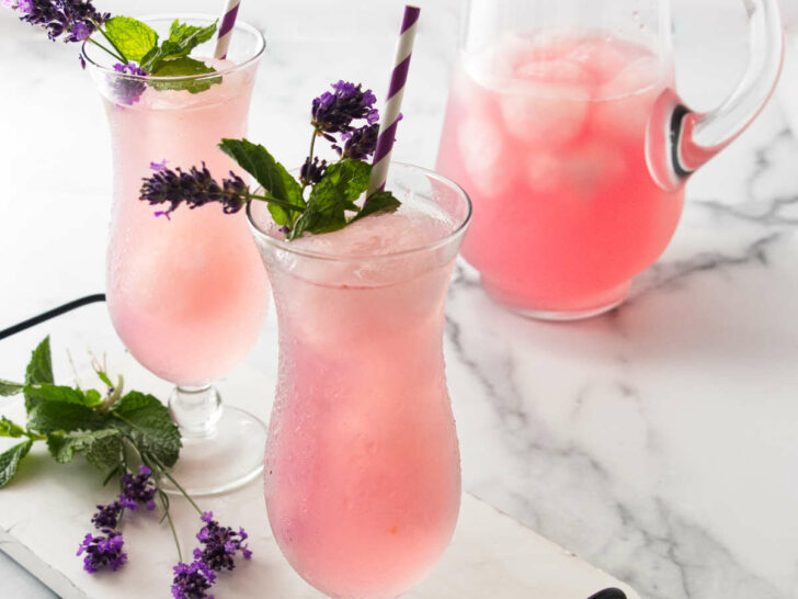 A clear glass pitcher and two tall glasses filled with a pink beverage sitting on a white tray with lavender blossoms.