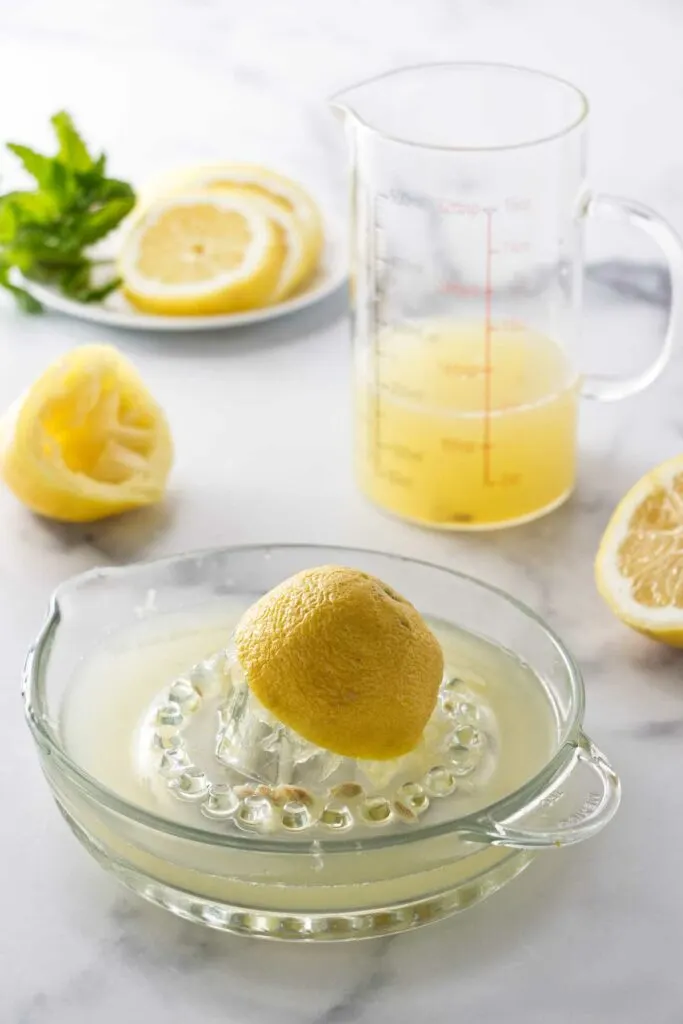 Half a lemon and lemon juice in a lemon juicer. A pitcher of lemon juice, lemons and a plate of lemon slices and mint.
