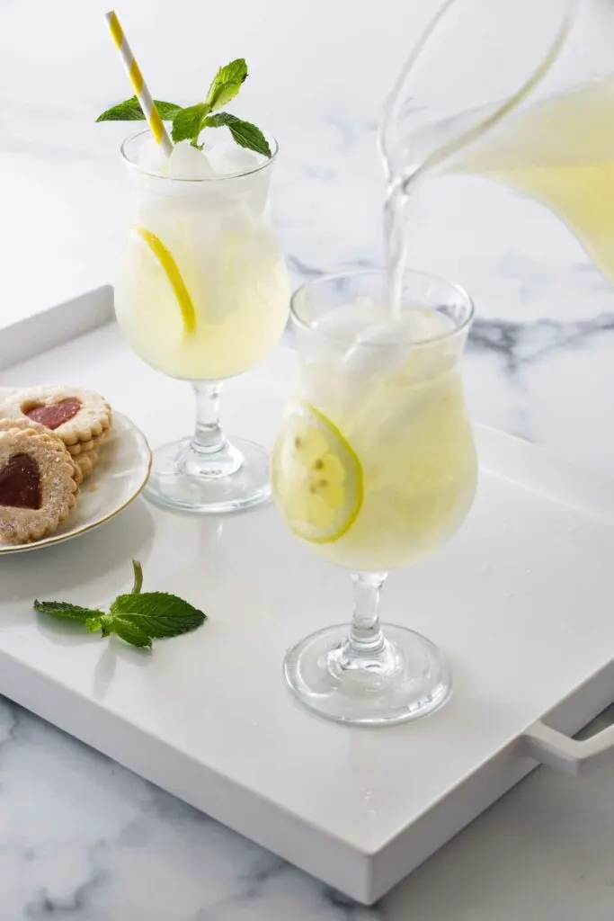 A serving tray with two glasses of old fashioned lemonade, and a plate of cookies. A pitcher of lemonade filling a glass with ice.