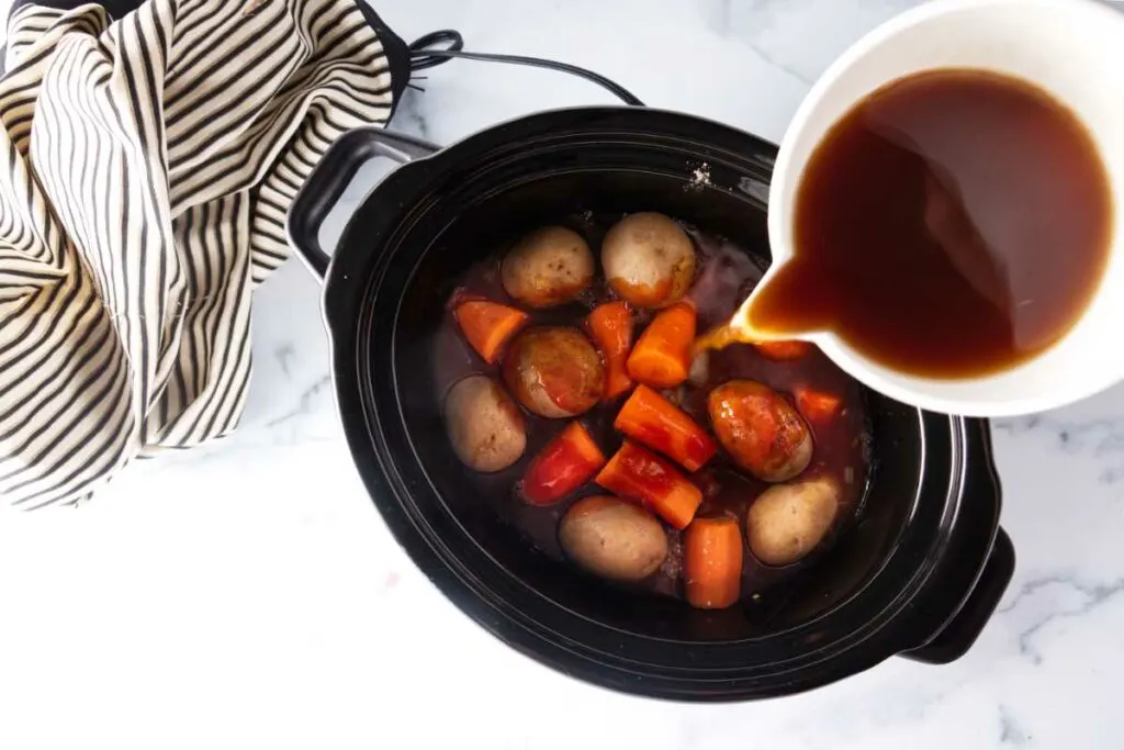 Pouring beef broth into a slow cooker bowl filled with beef, potatoes and carrots.