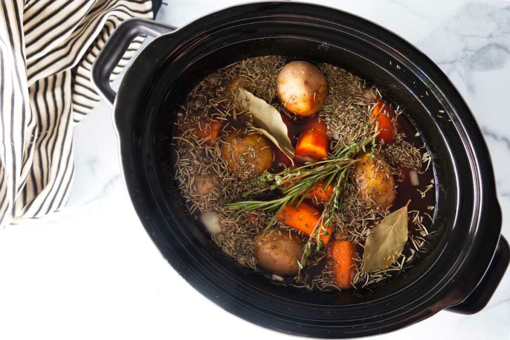 A slow cooker with vegetables and broth with dried herbs and bay leaves floating on top.