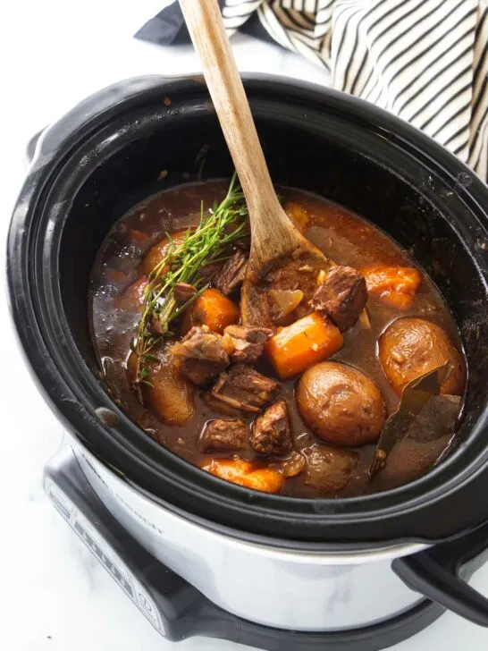 A wooden spoon in a slow cooker with carrots, potatoes and chunks of beef in a brown gravy sauce, garnished with fresh green herbs.