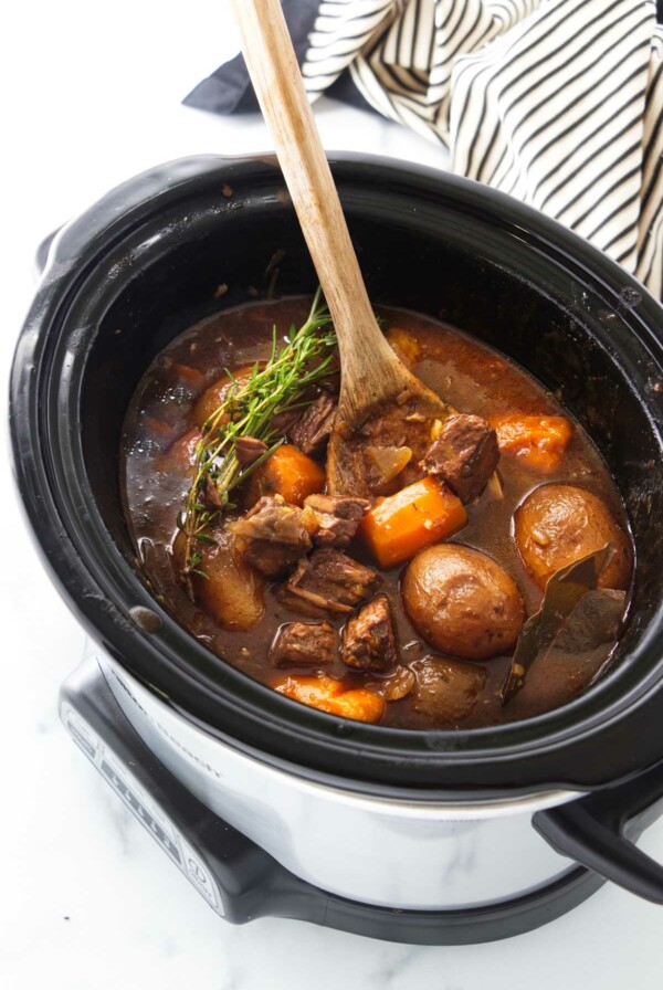 A wooden spoon in a slow cooker with carrots, potatoes and chunks of beef in a brown gravy sauce, garnished with fresh green herbs.