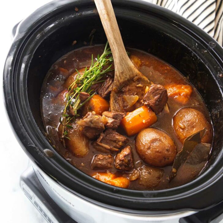 A wooden spoon in a slow cooker with carrots, potatoes and chunks of beef in a brown gravy sauce, garnished with fresh green herbs.