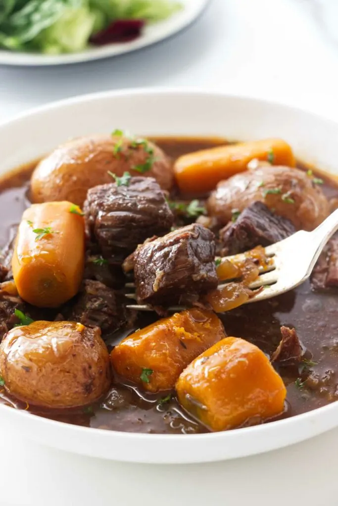 A dish of beef stew: carrots, potatoes, chunks of beef in a brown gravy sauce. A fork with a bite of feef.