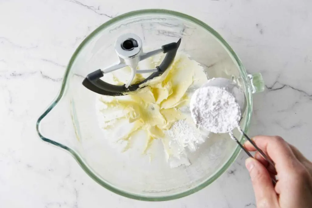 Adding powdered sugar to the creamed butter in a mixing bowl.