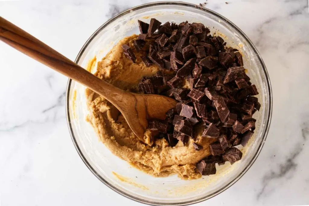 A bowl of cookie dough, chunks of chocolate and a wooden spoon.
