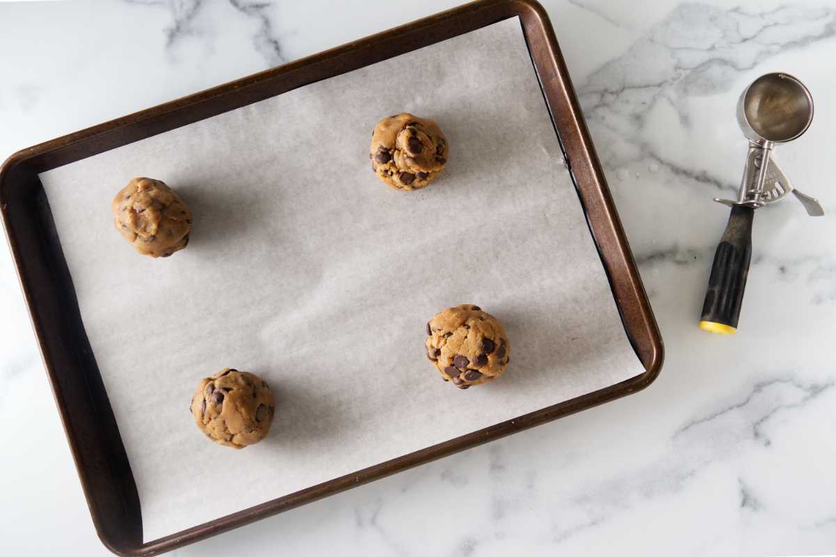 A parchment-lined baking sheet with four scoops of cookie dough and a large ice cream scoop next to the pan.