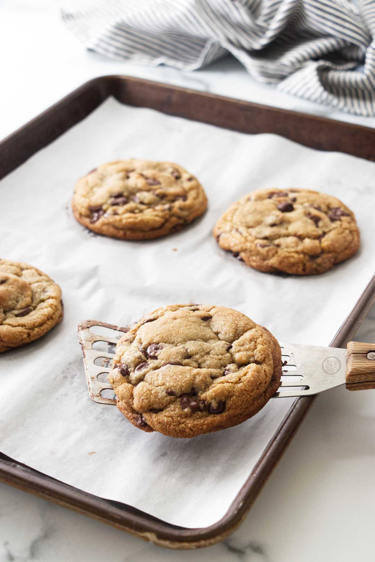 A parchment-lined baking pan with four giant cookies and a spatula lifting a cookie.