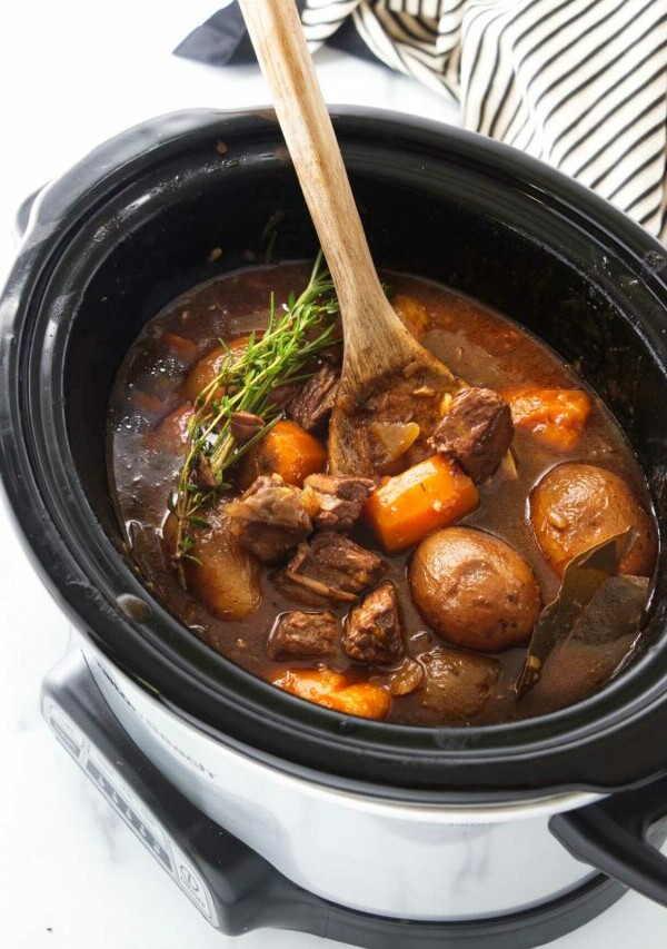 A wooden spoon in a slow cooker with carrots, potatoes and chunks of beef in a brown gravy sauce, garnished with fresh green herbs.