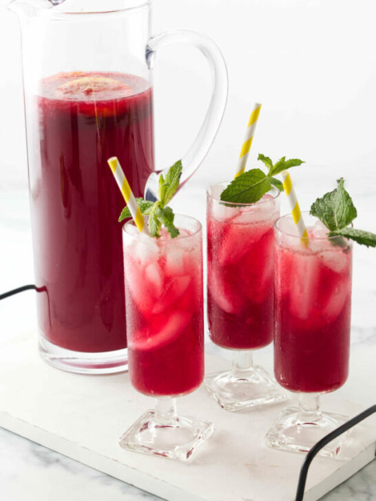 A serving tray with a pitcher filled with a red beverage and three glasses of red iced lemonade infused with hibiscus.