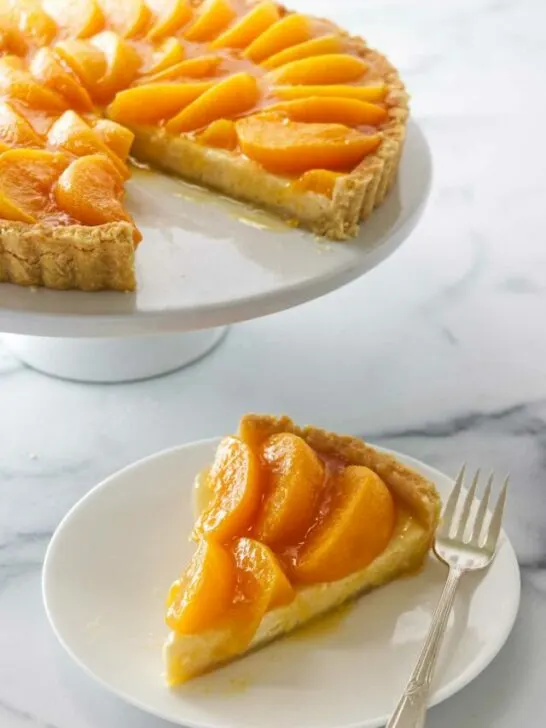 A peaches and cream tart on a cake pedestal in the background and a slice on a dessert plate with a fork in the foreground.